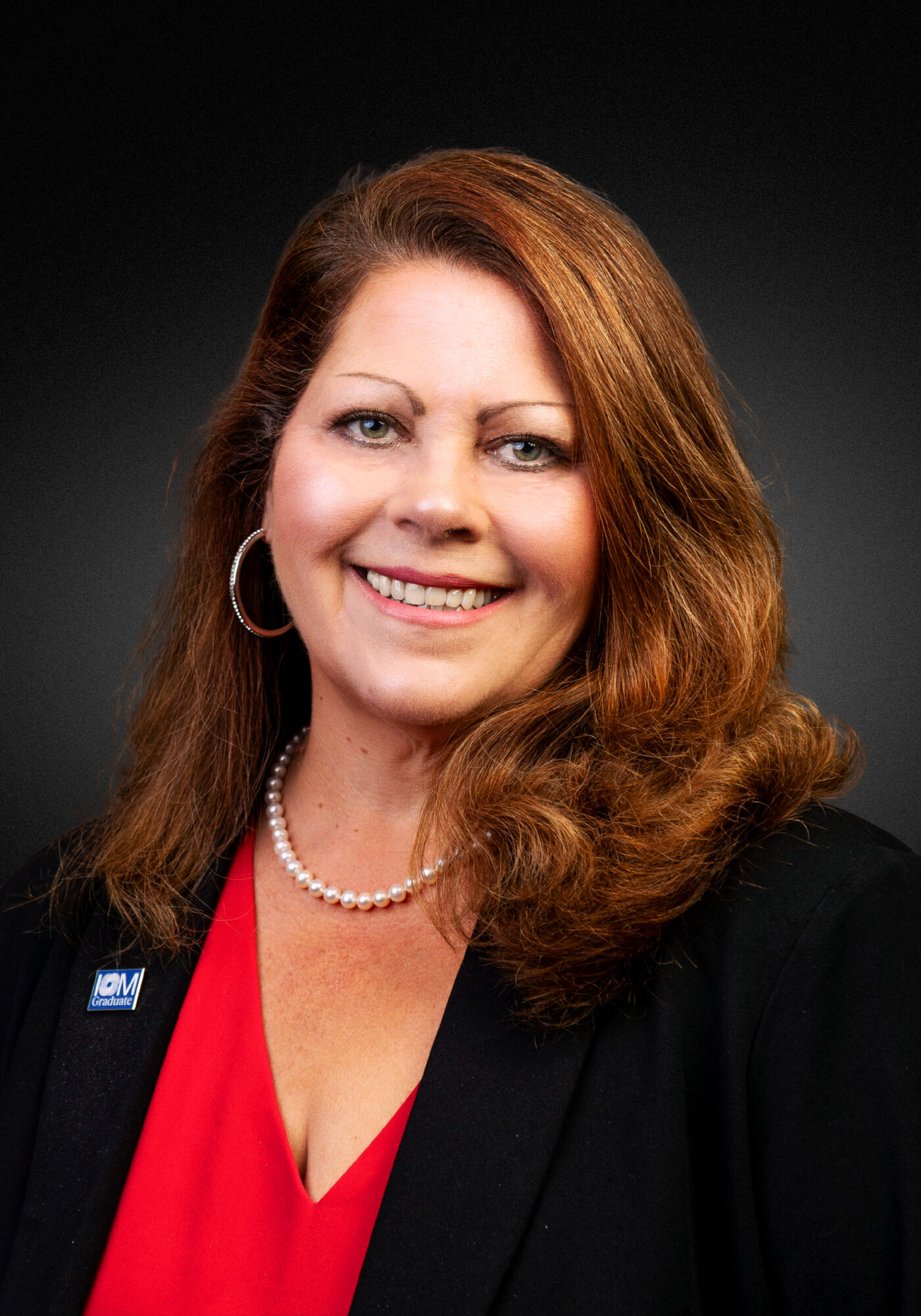 A woman with red-brown hair in a black suit jacket with a red shirt smiling at the camera